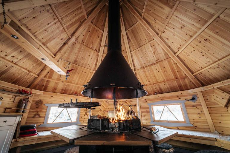 Puinen mökin sisätila, jossa suuri, keskellä oleva takka loimuaa. Seinillä koriste-esineitä ja ikkunoista näkyy talvinen maisema.