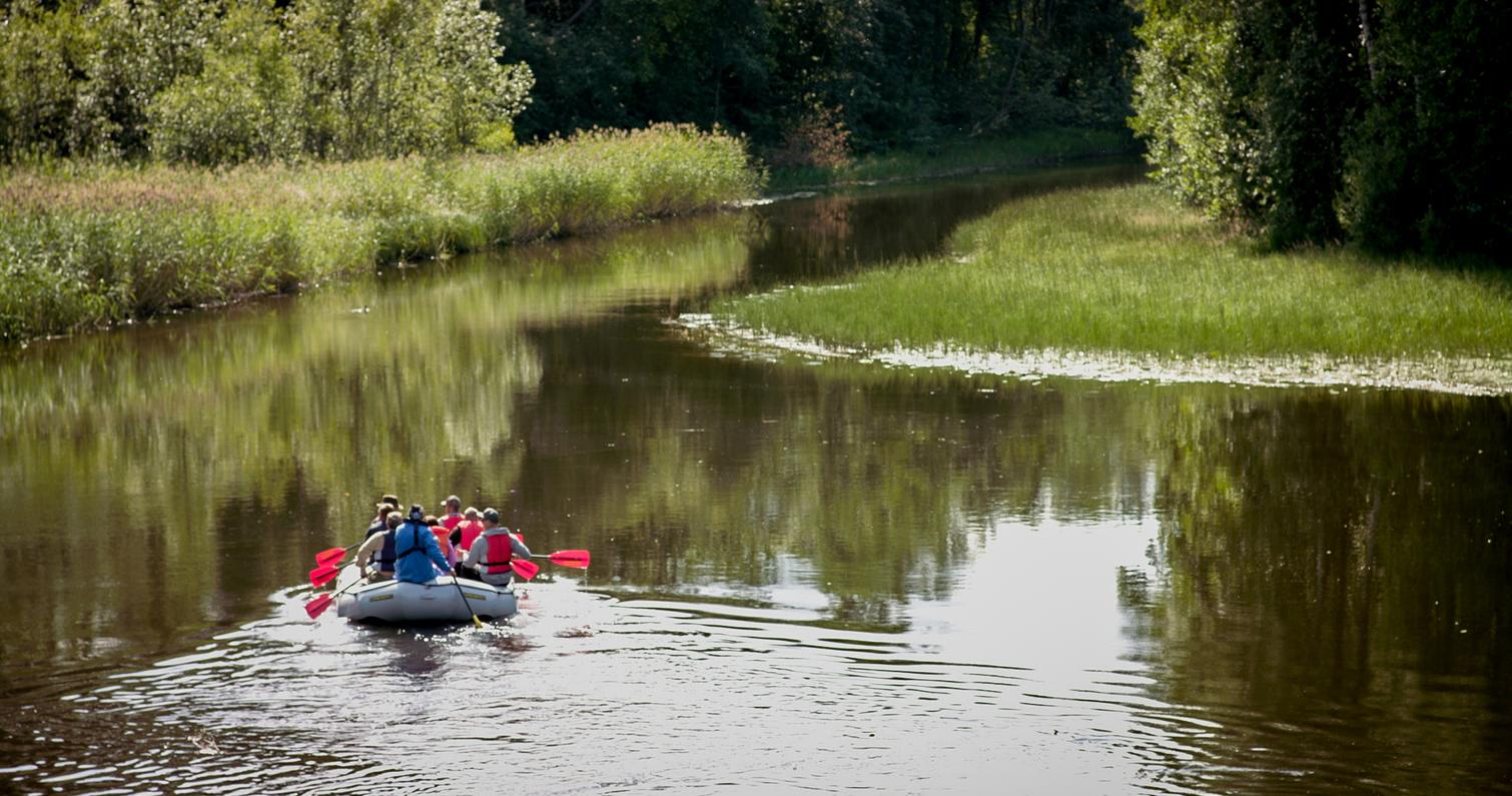 Ryhmä ihmisiä melontaretkellä kumiveneessä rauhallisella joella, jota ympäröivät vehreät rantakasvit ja metsät.
