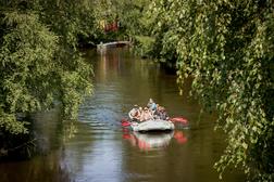 Ryhm&auml; ihmisi&auml; melomassa kumilautalla vehre&auml;n mets&auml;isen joen rauhallisessa maisemassa, puiden oksat kaartuvat veden ylle.