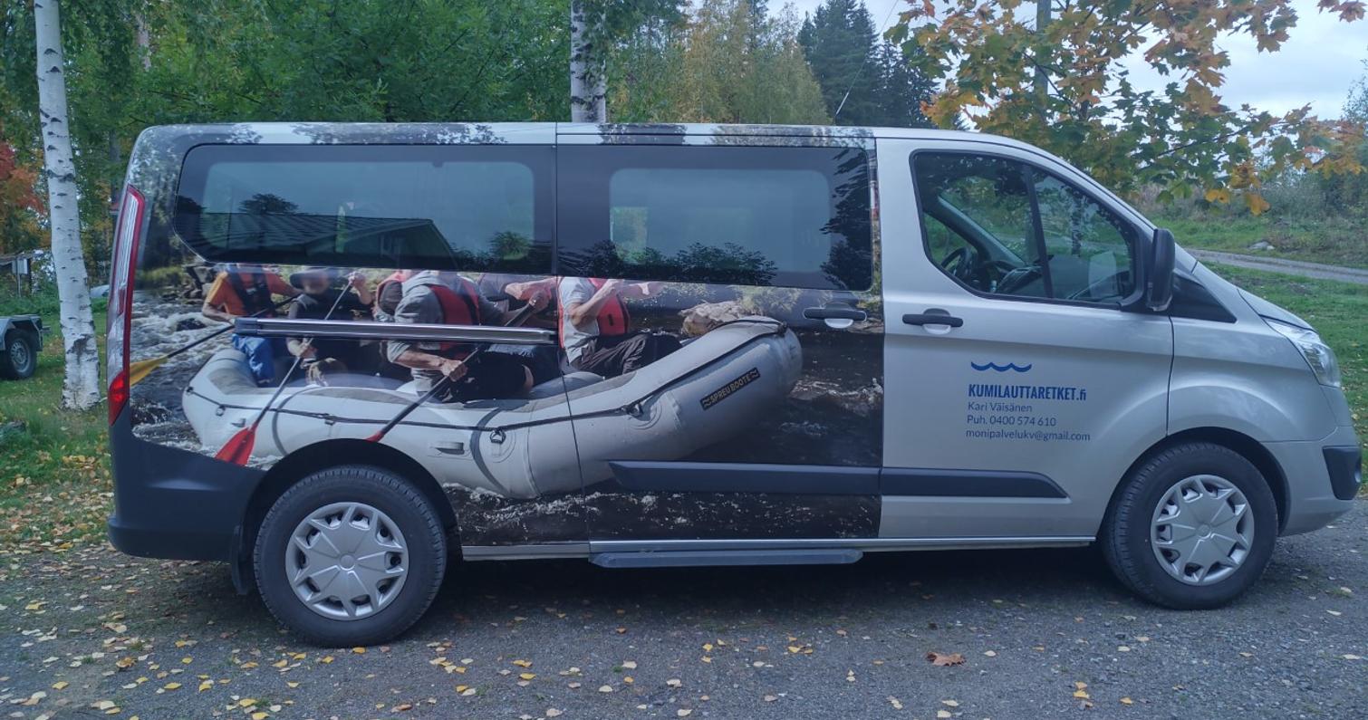 Mainoskuvioitu pakettiauto, jossa näkyy kumilauttailukuva ja yrityksen tiedot sivussa. Taustalla syksyinen metsä ja lehtipuut.