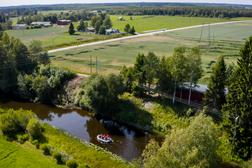 Ilmakuva joesta, jossa soutuvene kulkee rauhallista vesiv&auml;yl&auml;&auml;. Taustalla avarat niityt ja maalaistalot.