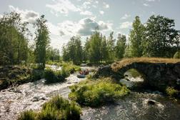 Rauhallinen joki virtaa kivisen kaarisillan ali, ympärillä vehreä metsämaisema ja kumilauttailijoita matkalla joen varrella.