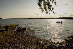 Ranta-alueella n&auml;kyy kivi&auml; ja nuotiopaikka, taustalla vesist&ouml; ja soutuvene, jossa on matkustajia auringonpaisteessa.