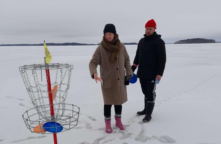 Kaksi henkilöä pelaamassa frisbeegolfia jäällä talvisessa maisemassa. Nainen heittää frisbeetä kohti koriin, ja mies katsoo vierestä.