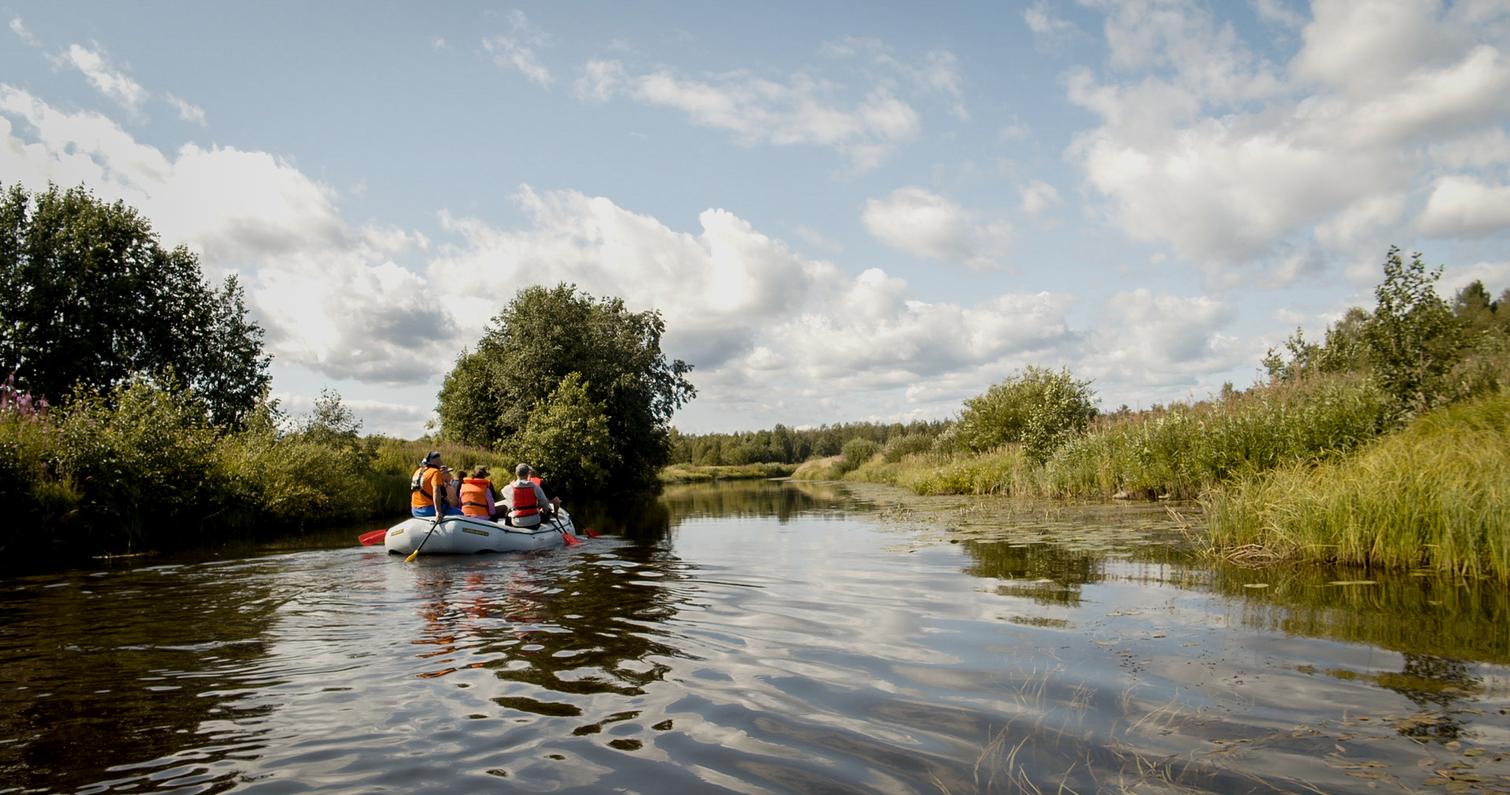 Ryhm&auml; ihmisi&auml; kelluu kumiveneell&auml; rauhallisella joella aurinkoisessa s&auml;&auml;ss&auml;, ymp&auml;rill&auml; vehreit&auml; puita ja kasvillisuutta.