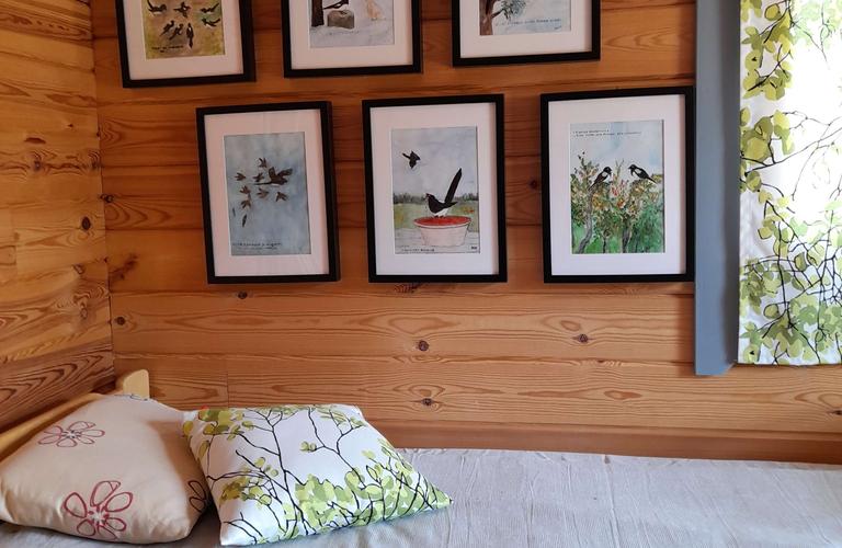 Puusein&auml;ll&auml; ripustettuja lintumaalauksia kehystettyin&auml; m&ouml;kin huoneessa. Sein&auml;&auml; vasten oleva s&auml;nky, jonka p&auml;&auml;ll&auml; on koristetyynyj&auml; ja luonnonl&auml;heiset verhot ikkunassa."