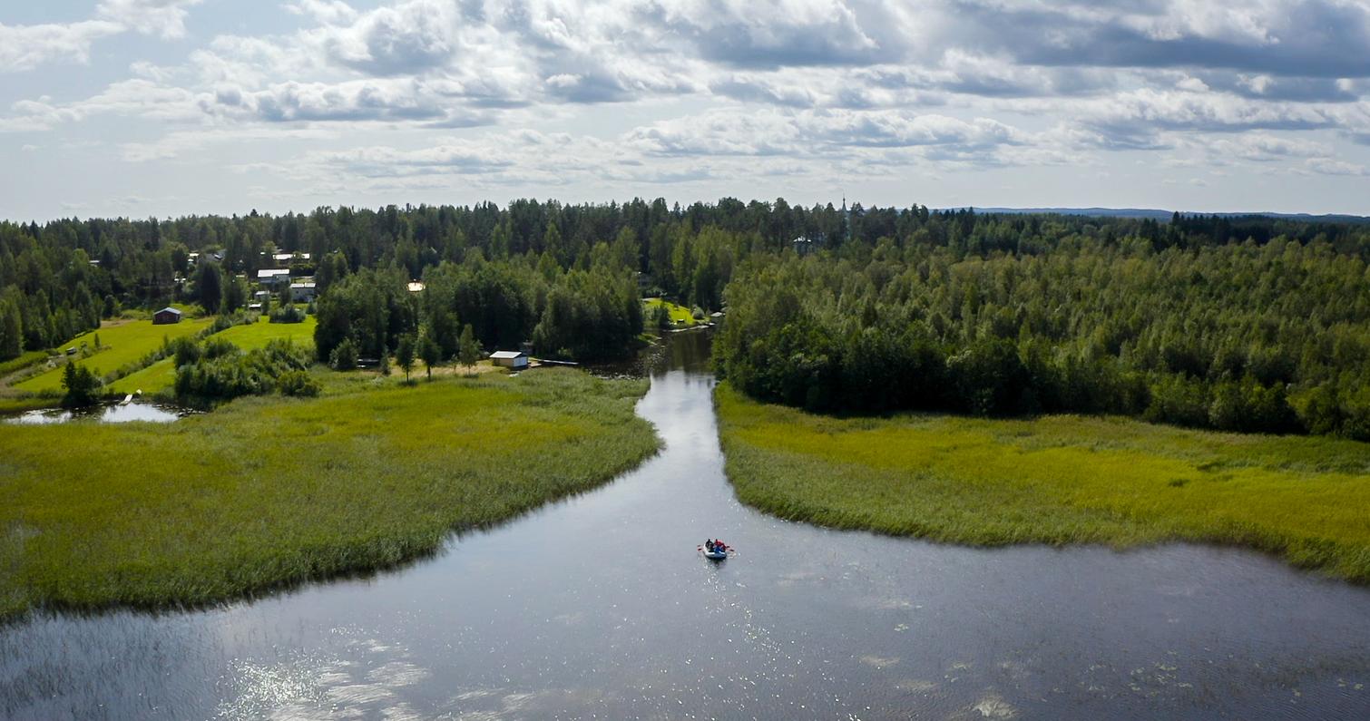 Ilmakuva rauhallisesta joesta, jota ympäröivät vehreät metsät ja niityt. Joella näkyy kumilauttailuryhmä keskellä luonnonmaisemaa.