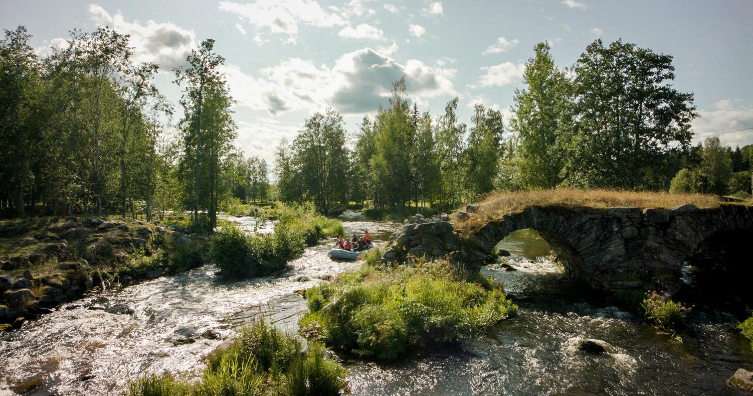 Rauhallinen joki virtaa kivisen kaarisillan ali, ympärillä vehreä metsämaisema ja kumilauttailijoita matkalla joen varrella.