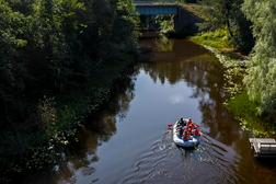 Ryhm&auml; ihmisi&auml; kelluu kumiveneell&auml; rauhallisessa joessa, jonka ymp&auml;rill&auml; on rehev&auml;&auml; kasvillisuutta ja heijastuksia vedess&auml;.