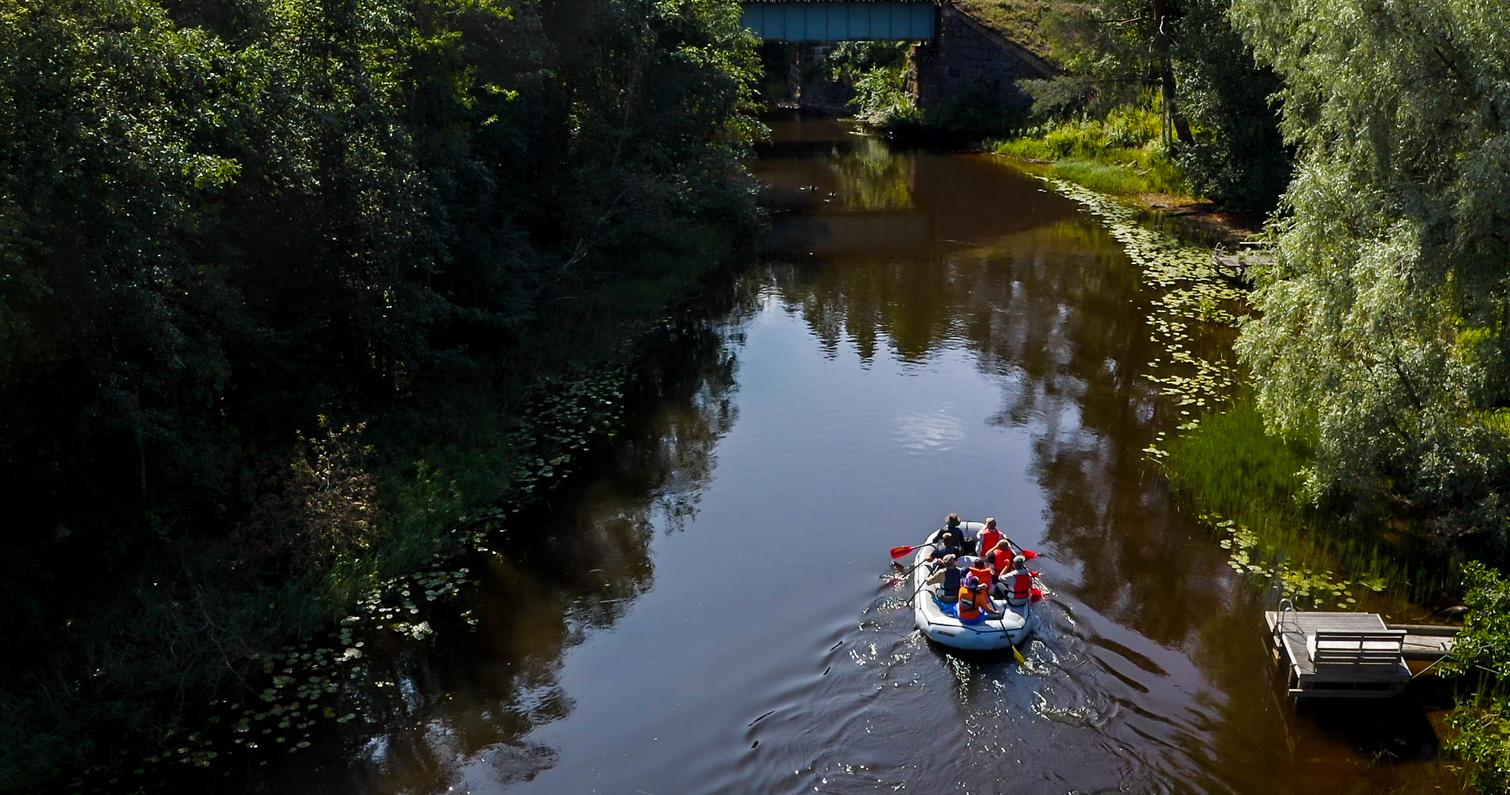 Ryhm&auml; ihmisi&auml; kelluu kumiveneell&auml; rauhallisessa joessa, jonka ymp&auml;rill&auml; on rehev&auml;&auml; kasvillisuutta ja heijastuksia vedess&auml;.
