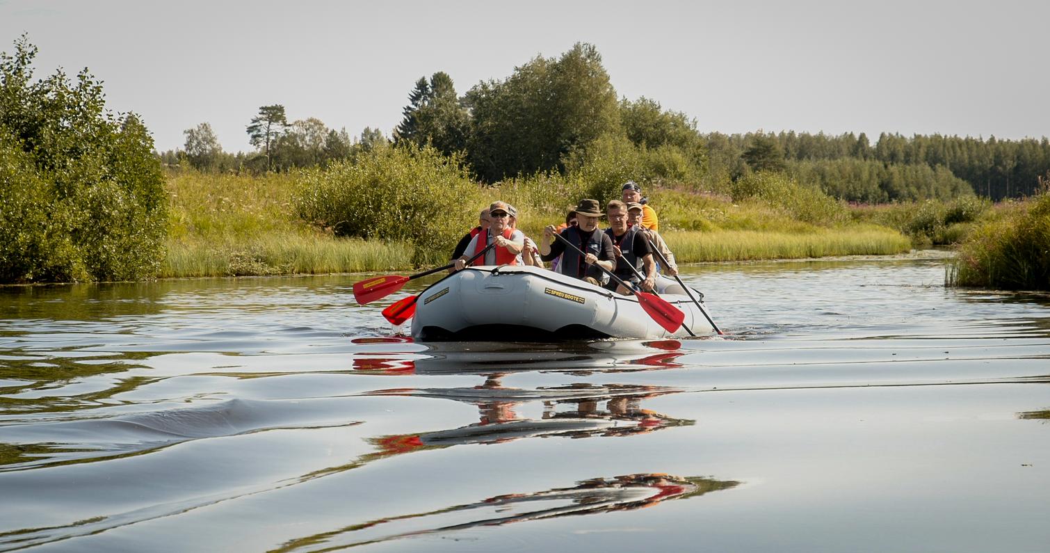 Ryhm&auml; ihmisi&auml; melomassa kumilautalla rauhallisella joella, ymp&auml;rill&auml;&auml;n vehre&auml; luonto ja kirkas taivas.