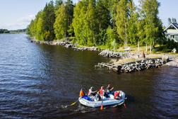 Ranta-alueella soutuvene kulkee järvellä, taustalla näkyy vihreitä puita ja rakennuksia auringonpaisteessa.