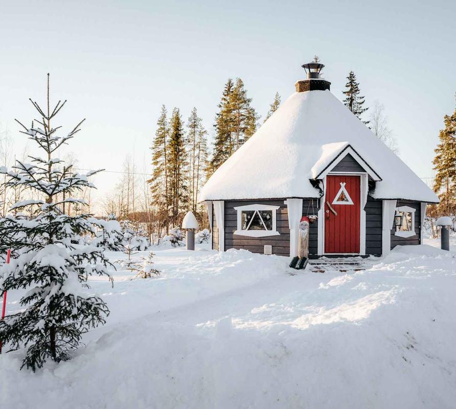 Talvinen mökki lumisessa maisemassa, jossa pieni kuusipuu etualalla ja punainen ovi korostaa mökin tunnelmallista ilmettä.