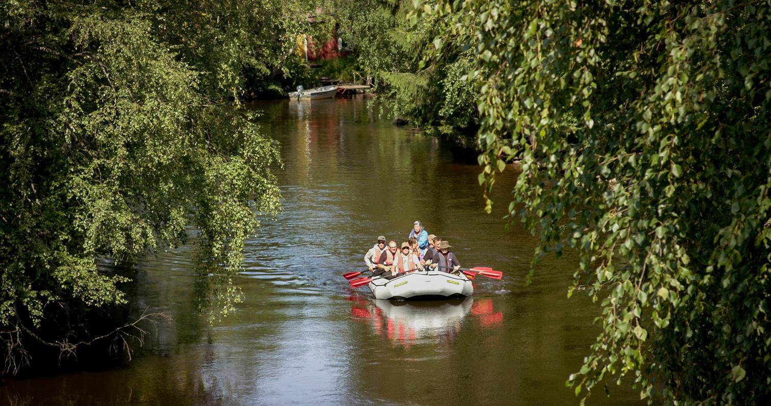 Ryhm&auml; ihmisi&auml; melomassa kumilautalla vehre&auml;n mets&auml;isen joen rauhallisessa maisemassa, puiden oksat kaartuvat veden ylle.