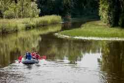 Ryhm&auml; ihmisi&auml; melontaretkell&auml; kumiveneess&auml; rauhallisella joella, jota ymp&auml;r&ouml;iv&auml;t vehre&auml;t rantakasvit ja mets&auml;t.