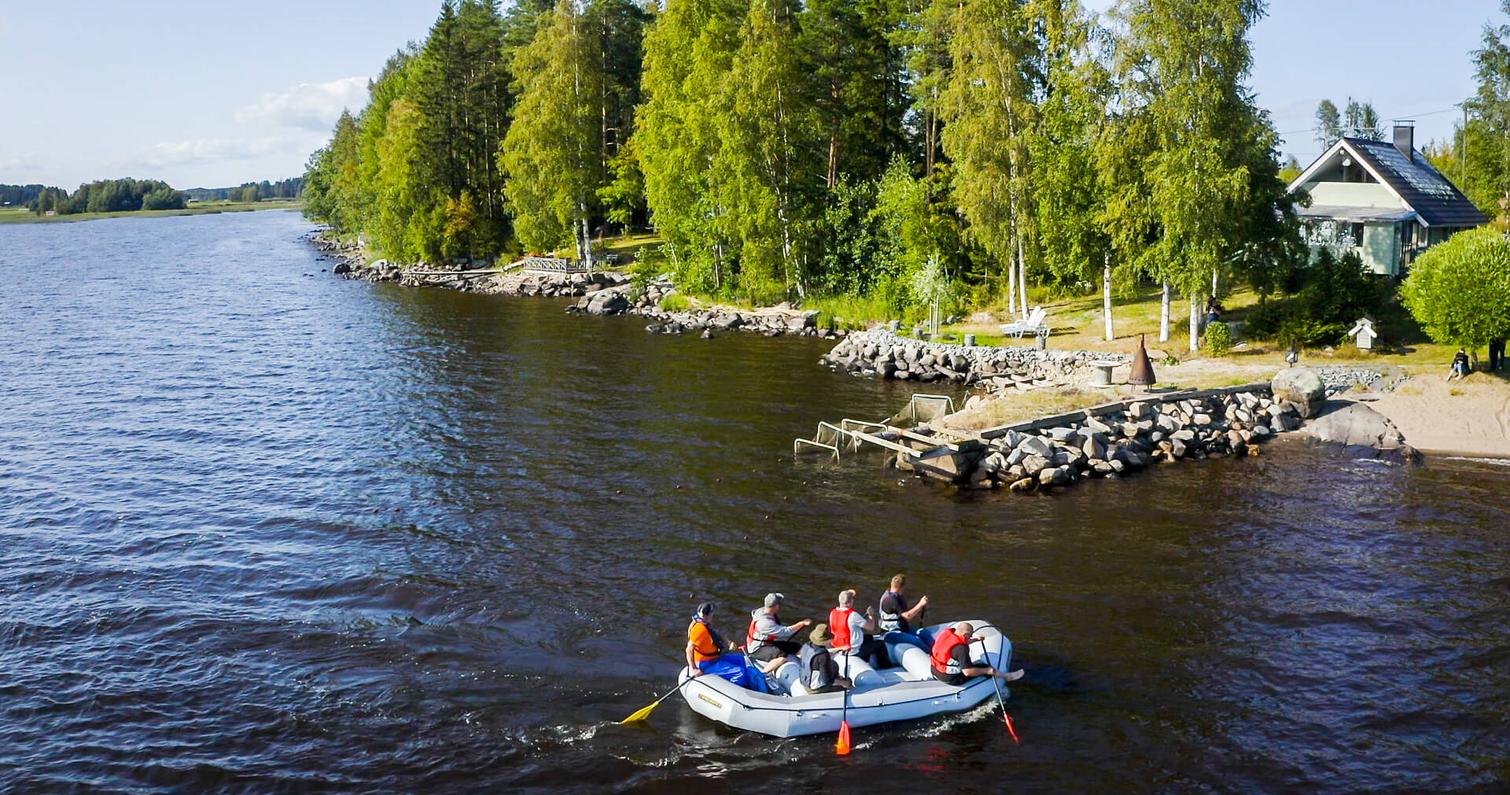Ranta-alueella soutuvene kulkee järvellä, taustalla näkyy vihreitä puita ja rakennuksia auringonpaisteessa.