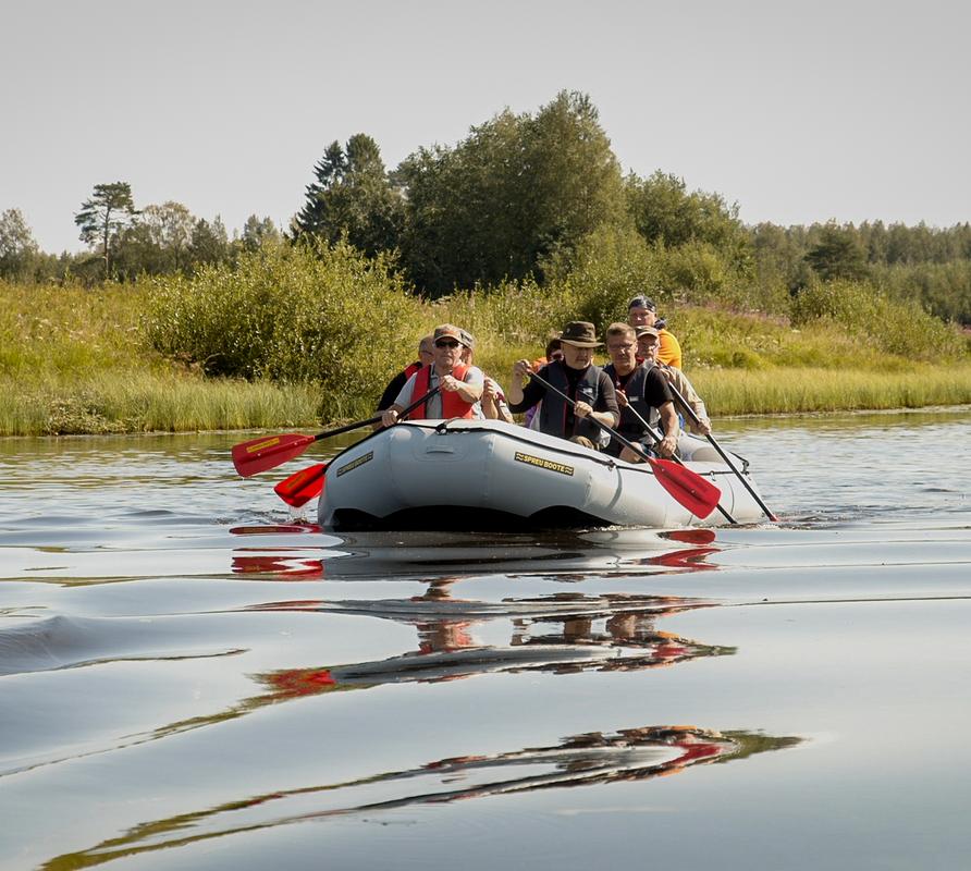 Ryhm&auml; ihmisi&auml; melomassa kumilautalla rauhallisella joella, ymp&auml;rill&auml;&auml;n vehre&auml; luonto ja kirkas taivas.