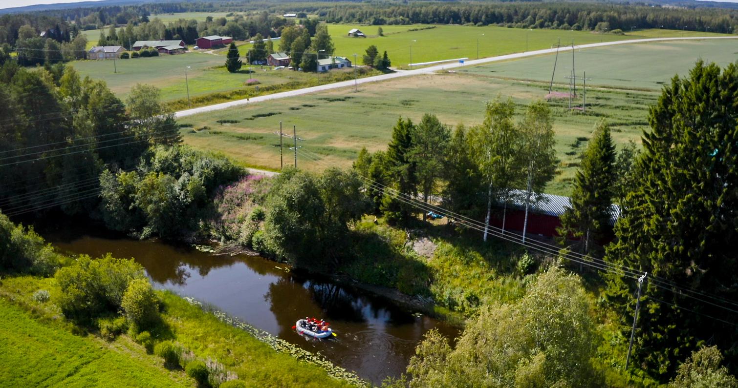 Ilmakuva joesta, jossa soutuvene kulkee rauhallista vesiväylää. Taustalla avarat niityt ja maalaistalot.