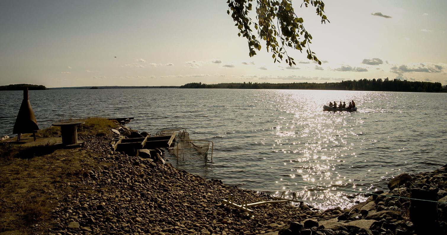 Ranta-alueella näkyy kiviä ja nuotiopaikka, taustalla vesistö ja soutuvene, jossa on matkustajia auringonpaisteessa.