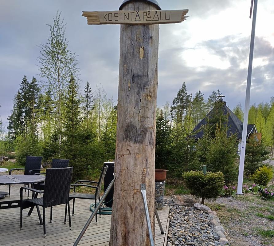 Puupylv&auml;s, jossa on kyltti 'Kosintapaalu', seisoo terassilla luonnon ymp&auml;r&ouml;im&auml;n&auml;. Taustalla n&auml;kyy tuoleja ja p&ouml;yt&auml; sek&auml; m&ouml;kki vehre&auml;ss&auml; maisemassa.
